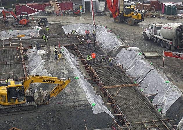 an excavator sites on dirt with a trench that has wide concrete forms with rebar and concrete pump nozzle leading into it.